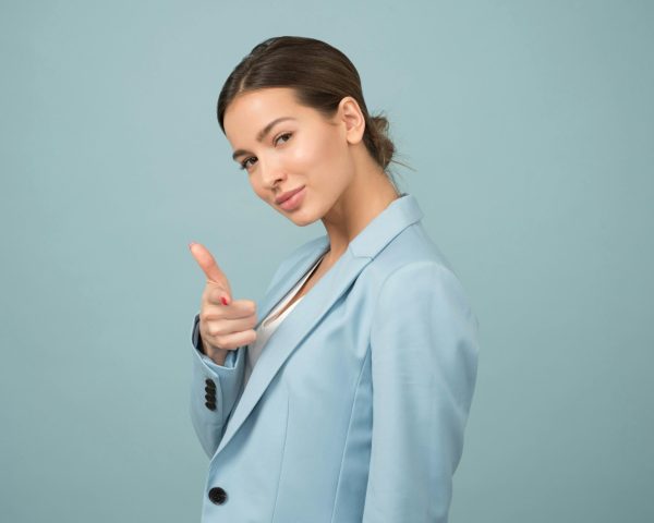 A young woman in a blue suit exhibits confidence with a relaxed pose against a blue background.